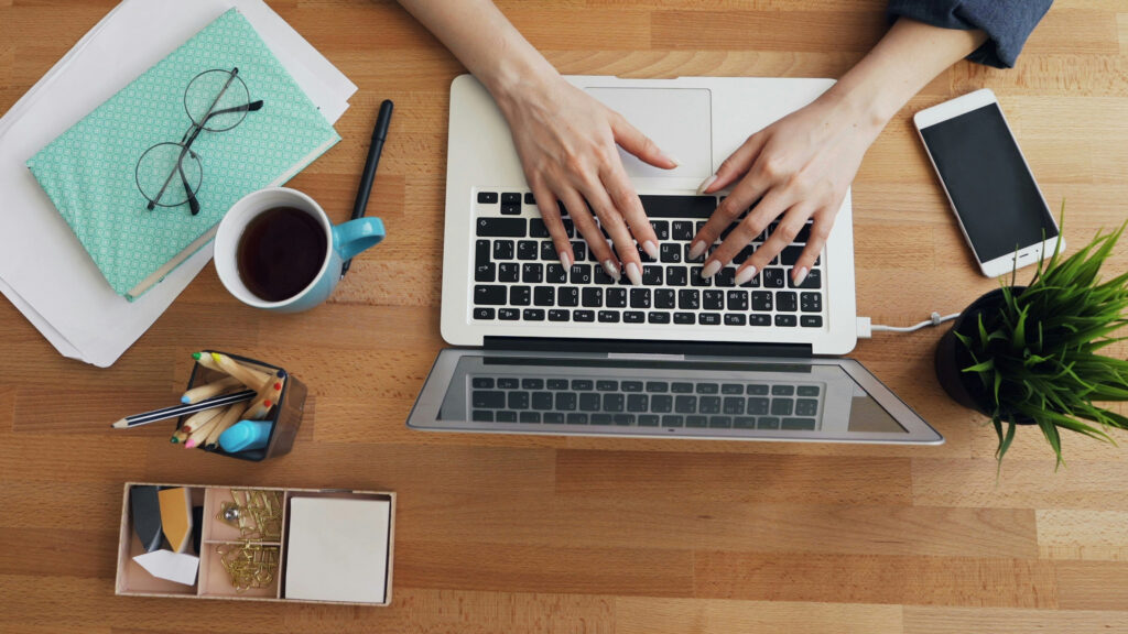 Person arbeitet am Laptop an einem aufgeräumten Holzschreibtisch mit Kaffee und Notizbuch